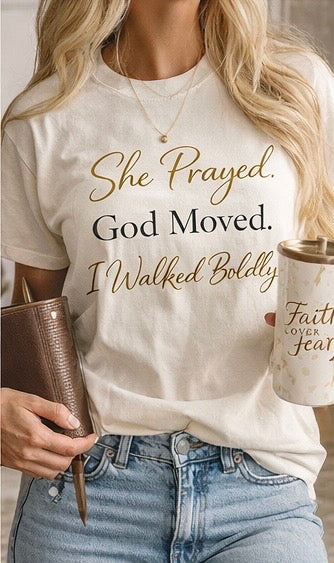 Person wearing a white t-shirt with text She Prayed, God Moved... I walked Boldly holding a coffee cup and book.
