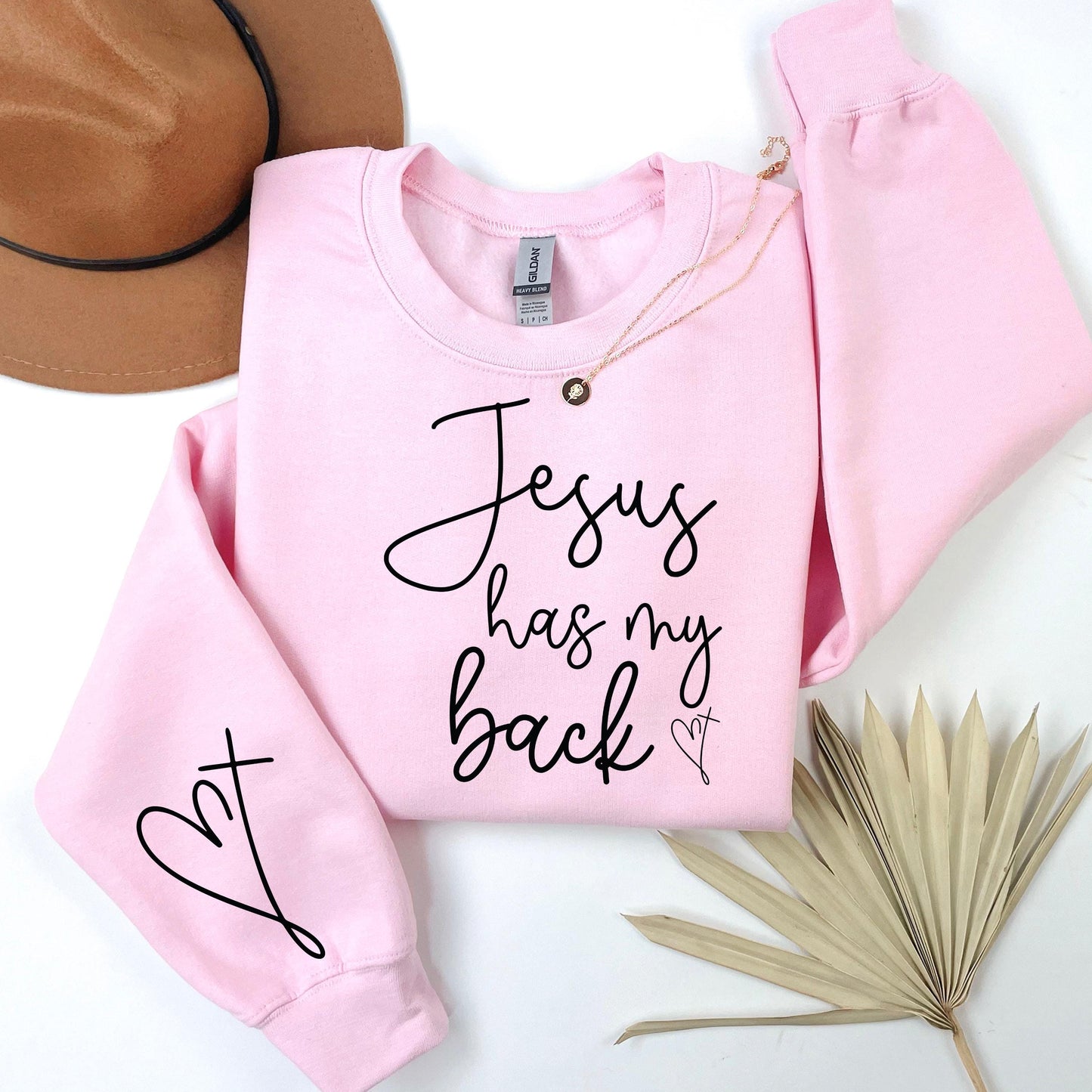 Pink sweatshirt with 'Jesus has my back' text on a white surface with a hat and dried leaves.