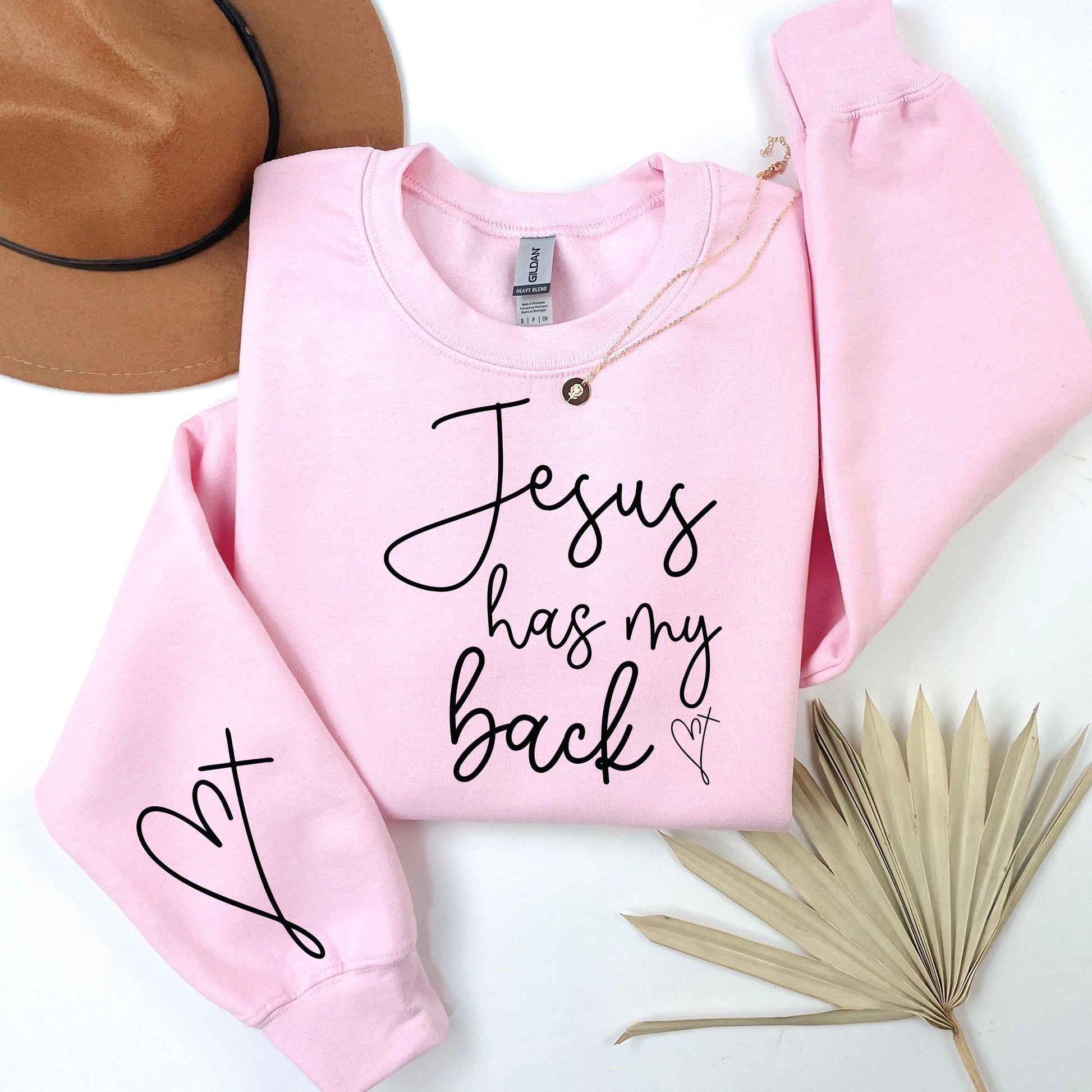 Pink sweatshirt with 'Jesus has my back' text on a white surface with a hat and dried leaves.